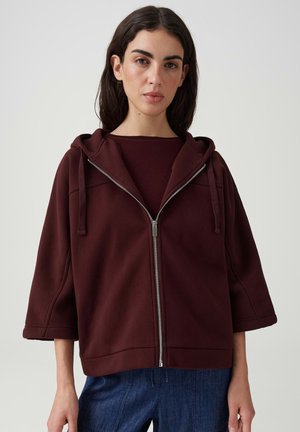 Frau mit dunklen Haaren, die einen bordeauxfarbenen Kapuzenpullover mit Dreiviertelärmeln und Reißverschluss sowie blaue Jeans vor einem einfarbig hellen Hintergrund trägt.