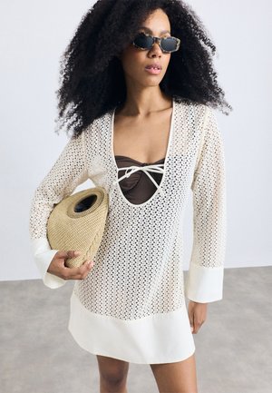 Donna con capelli ricci che indossa occhiali da sole tartarugati, un abito in cotone bianco con una maglietta marrone sotto, mentre tiene in mano una borsa rotonda di paglia.