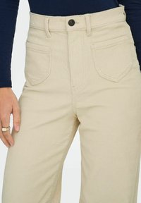 Pantalons en velours côtelé beige à taille haute, avec poches avant, une fermeture à bouton visible et une texture côtelée verticale.