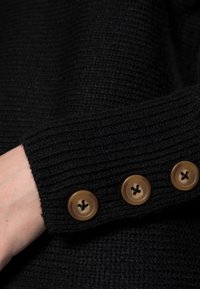 Schwarzer gestrickter Pulloverbund mit geripptem Muster und drei braunen Knöpfen mit einem Fadenmuster.