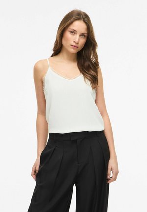Femme aux longs cheveux bruns portant un caraco blanc orné de dentelle et un pantalon noir plissé taille haute sur fond uni.
