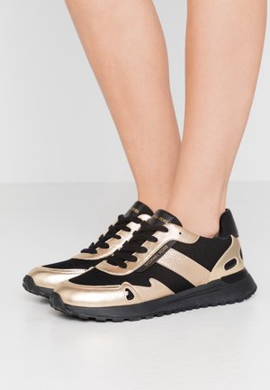 Nogi w czarnych i metalicznie złotych butach sportowych Michael Kors z czarnymi sznurowadłami, stojące na jasno szarej podłodze.