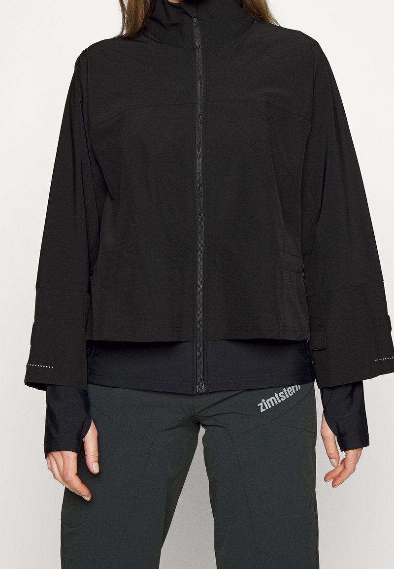 Czarna kurtka z wysokim kołnierzem, zamkiem z przodu, szerokimi rękawami i odblaskowymi akcentami. Gładki materiał, minimalistyczny design, zestawiona z ciemnymi spodniami.