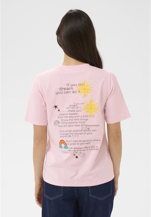 Femme aux longs cheveux foncés portant un t-shirt rose avec un texte inspirant, des soleils, des étoiles et un arc-en-ciel au dos.