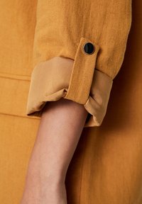 Manche roulée de couleur moutarde avec un bouton noir sur l'avant-bras d'une personne, sur un fond de tissu assorti.