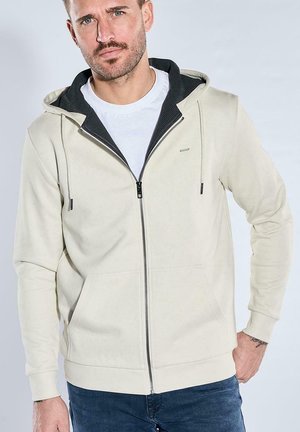 Mann mit leichtem Bart, der einen beigen Zip-Hoodie über weißem Shirt und blauen Jeans trägt, steht mit einer Hand in der Tasche vor einfarbigem Hintergrund.