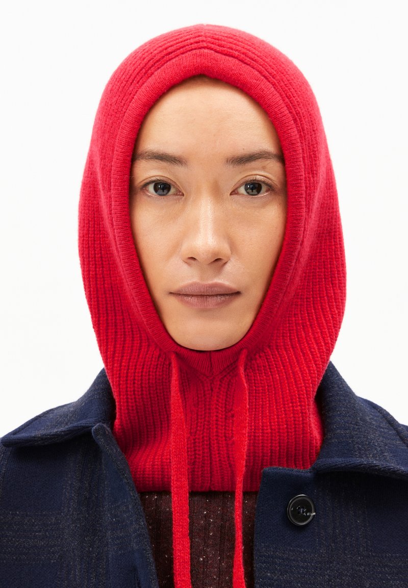 Rote gestrickte Kapuze mit geripptem Muster, ausgestattet mit einer Kordel am Hals. Getragen über einem dunkelblauen Mantel, mit einem braun gemusterten Pullover darunter.