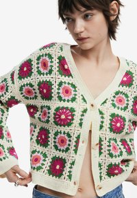 Virkat kort sweatjacka med blommönster i rosa, grön och krämfärgad, med stora knappar och öppen frontdesign. Texturerad, lätt tyg.