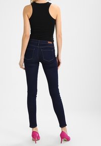 Jean skinny en denim foncé, avec poches arrière et un écusson en cuir sur la ceinture. Associé à un débardeur noir et des talons roses.