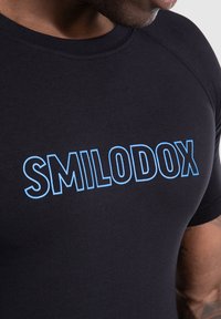 Camiseta negra ajustada hecha de una tela elástica con "SMILODOX" impreso en letras bold de color azul claro en el pecho. Manga corta, cuello redondo.