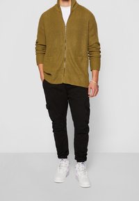 Olijfgroene gebreide cardigan met een omslagkraag, ritssluiting, twee steekzakken aan de voorkant, gecombineerd met zwarte cargobroeken en witte sneakers.