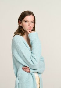 Light blue, oversized cardigan made of soft, textured knit fabric. Features wide sleeves and deep V-neckline with pockets.