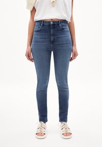 Jeans ajustados en denim azul oscuro con cintura alta, cinco bolsillos y un desvanecimiento mínimo; combinados con sandalias blancas con cordones.