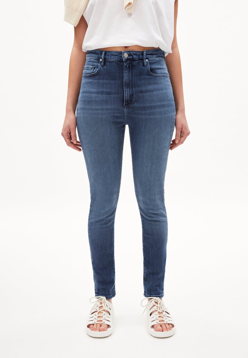 Jeans ajustados en denim azul oscuro con cintura alta, cinco bolsillos y un desvanecimiento mínimo; combinados con sandalias blancas con cordones.