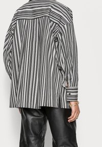 Chemise oversize à rayures noires et blanches, manches longues, poignets à boutons, et ourlet arrondi. Fabriquée en tissu léger avec une texture visible.