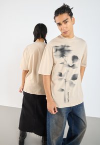 Camiseta beige de manga corta con un diseño floral negro y texto en el frente, combinada con unos jeans oscurecidos y una falda negra.