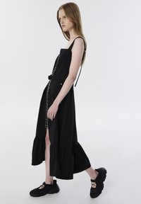 Zwarte midi-jurk met een uitlopende zoom, voorzien van een rits aan de voorkant en verstelbare bandjes. Gecombineerd met stevige zwarte sneakers.