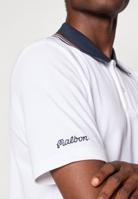 Valge polo särk tumesinise kaelusega, millel on oranžid triibud. Vasakul varrukal on tumesinise värvusega broderitud "Malbon". Puuvillane materjal, lühikesed varrukad.