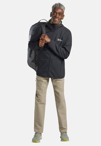 Veste softshell noire avec fermeture éclair à l'avant, portant un pantalon beige clair, des baskets grises et portant un sac à dos noir avec des accents réfléchissants.