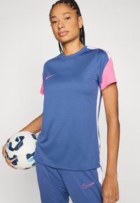 Αθλητική μπλούζα Nike σε μπλε με ροζ κοντά μανίκια και λευκές λεπτομέρειες. Φοριέται με παντελόνια που ταιριάζουν, κρατώντας μια μπάλα ποδοσφαίρου. Υφή υφάσματος.