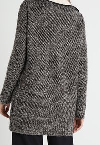 Överdimensionerad lång cardigan i en grå och svart texturerad stickning, med en bred halsringning och kontrasterande kant längs kanterna.