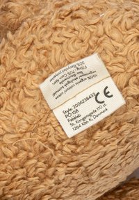 Beige, strukturierte Hausschuhe aus 30 % recyceltem Polyester und 70 % biologischer Baumwolle, mit einem Stoffetikett, das Pflegehinweise enthält.