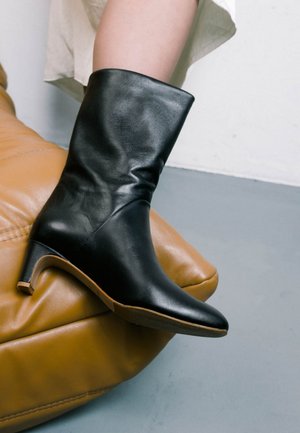 Stivale alla caviglia in pelle nera con punta affusolata e un tacco modesto, caratterizzato da una texture liscia e una leggera piega, posizionato su una superficie in pelle marrone.