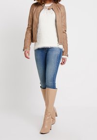 Brun läderjacka, vit fluffig tröja, blå smala jeans och beige knähöga klackstövlar. Modellen står med en hand på höften.