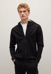 Felpa nera con zip e cappuccio regolabile, in tessuto morbido, due tasche frontali e design aderente. Presenta una superficie liscia e dettagli minimi.