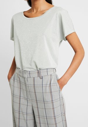 Vrouw draagt een lichtzilver glinsterende top met korte mouwen, ingestopt in hoge taille lichtgrijze geruite broek, met handen in de zakken.