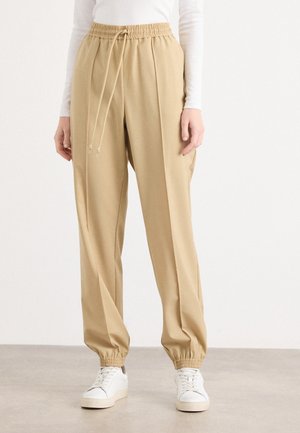 Person wearing beige drawstring jogger pants with elastic cuffs and white sneakers, standing on a gray floor against a plain background.