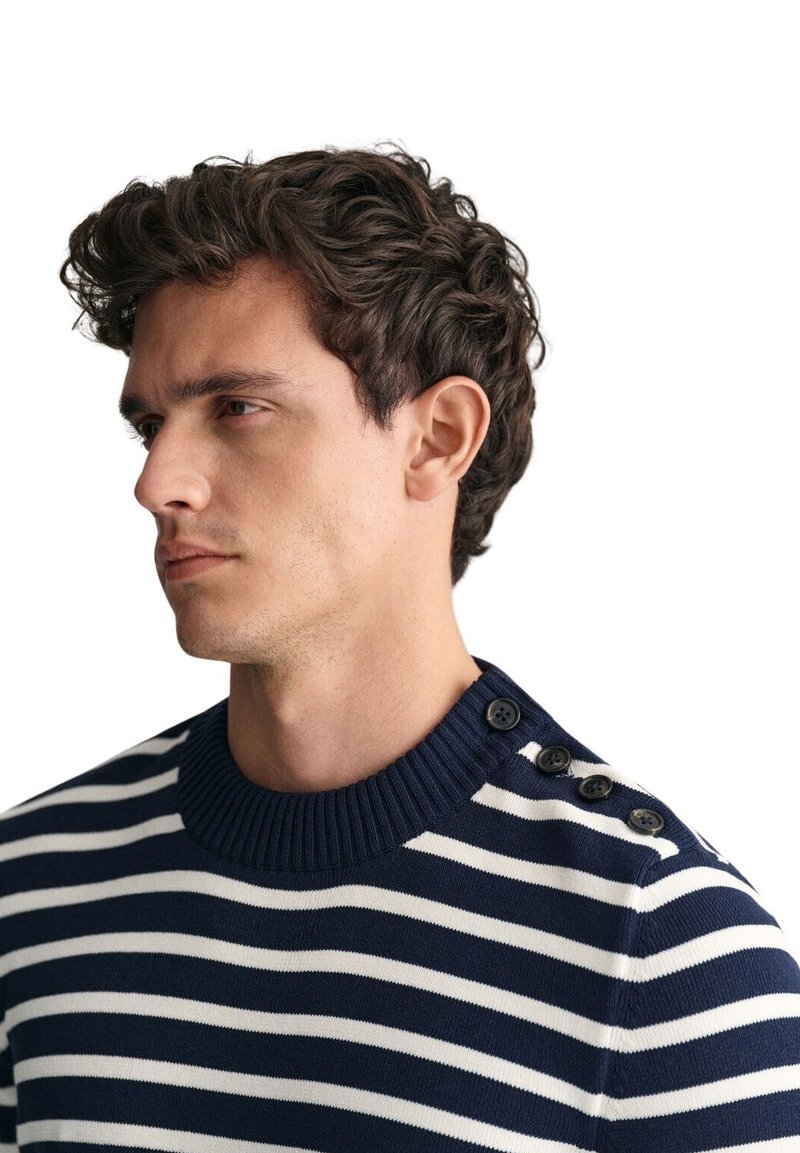 Joven con cabello rizado y castaño que lleva un suéter de rayas azul marino y blanco con cuatro botones negros en el hombro, mirando hacia la izquierda.