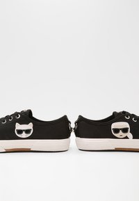 Musta matalaankengät, jotka kohtaavat toisensa, kummassakin valkoiset pohjat ja ulkopuolella kissan ja aurinkolaseja käyttävän ihmisen kuvat.