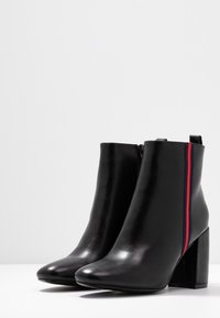 Bottes à cheville en cuir noir avec un bout carré et un talon carré. Dotées d'une fermeture éclair et d'une bande d'accent rouge et bleue sur le côté.
