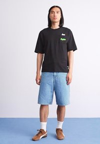 Black cotton t-shirt with a green logo on the pocket, paired with blue denim shorts. Brown sneakers and white crew socks complete the look.