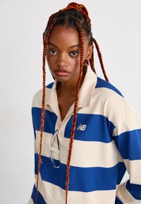 Jeune personne avec de longues tresses rouges portant une chemise rayée bleue et blanche avec des lunettes suspendues au col, regardant directement devant.