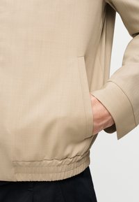 Veste beige avec poche latérale et taille élastique, main reposant dans la poche, associée à un pantalon noir sur un fond uni.