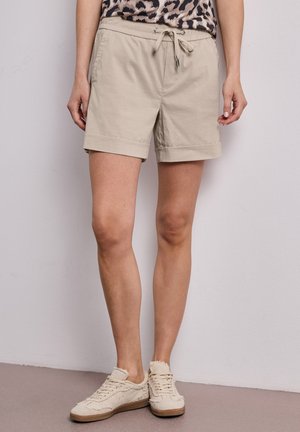 Kobieta w beżowych szortach ze sznurkiem, topie w panterkę oraz postrzępionych kremowych trampkach, stojąca przy gładkiej białej ścianie.