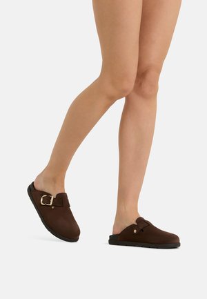Glatt rasierte Beine tragen braune Slip-On-Clogs mit schwarzen Sohlen und goldener Schnallendetail, die auf einem schlichten hellgrauen Hintergrund gehen.