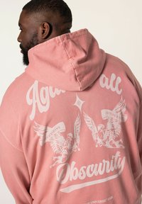 Sudadera rosa con un gran gráfico blanco en la parte posterior que presenta dos figuras aladas y el texto "Contra toda Oscuridad" y "Engañoso".
