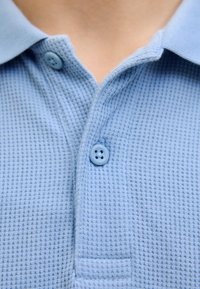 Hellblauses Polohemd aus strukturiertem Waffelstrickstoff, mit einer Knopfleiste mit zwei Knöpfen und einem weichen Kragen.
