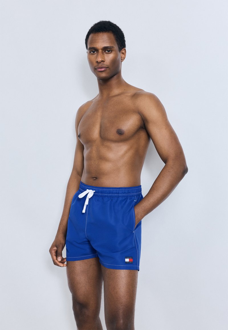 Μπλε μαγιό σορτς με ελαστική μέση και λευκό κορδόνι, πλαϊνές τσέπες και μικρό λογότυπο στην αριστερή πλευρά.