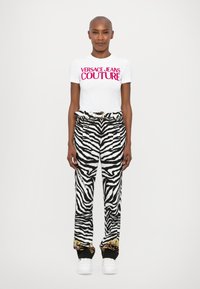 Baltā t-krekls ar tekstu "VERSACE JEANS COUTURE" rozā krāsā, saskaņots ar melnbaltām zebra svītrainām biksēm ar zelta akcentu apakšmalu. Baltas sporta kedas.