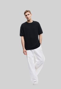 Zwarte oversized t-shirt met korte mouwen, gecombineerd met losse witte broeken en witte sneakers tegen een grijze achtergrond.