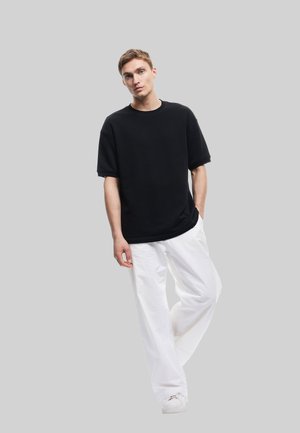 Svart oversized t-shirt med korta ärmar, kombinerad med löst sittande vita byxor och vita sneakers mot en grå bakgrund.