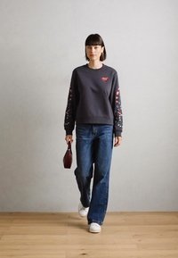 Felpa grigia scura con fantasie floreali colorate sulle maniche, abbinata a jeans a gamba larga blu e sneakers bianche. Tiene una piccola borsa bordeaux.