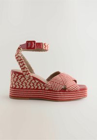 Sandales compensées rouges avec un dessus tissé présentant un motif en chevrons, une bride de cheville réglable et une zone des orteils lisse et brillante.