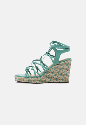 Turquoise wedge sandaal met een gevlochten stro-zool, voorzien van crisscross bandjes en een open teenontwerp, wat de ademend vermogen en stijl versterkt.