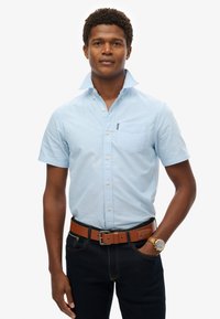 Jasnoniebieska koszula krótko rękawowa z kołnierzykiem zapinanym na guziki, z kieszenią na piersi. Noszona z ciemnymi dżinsami i brązowym skórzanym paskiem.