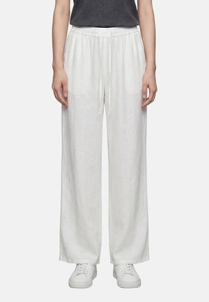 Personne portant un pantalon blanc ample à cordon, des baskets blanches et un haut gris foncé, debout devant un fond uni.